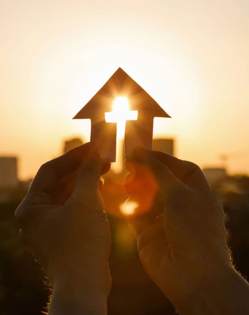 hands holding paper house with crosss removed and sunrays streaming through