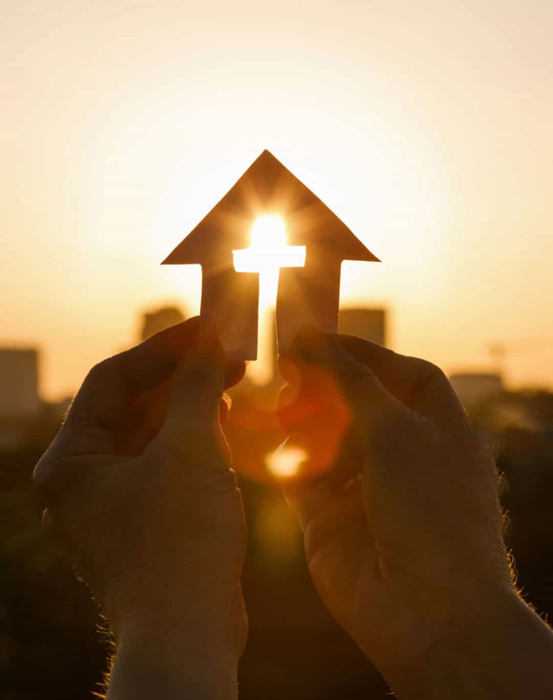 hands holding paper house with crosss removed and sunrays streaming through